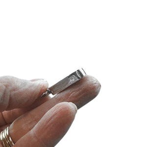 May include: A silver-colored pendant held in a hand. The pendant has a rectangular shape with a rounded edge. The hand has multiple gold rings on one finger. The background is white.