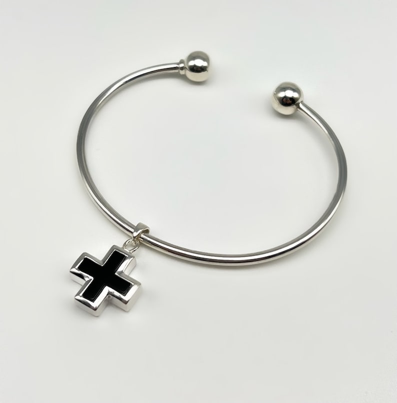 May include: A silver bangle bracelet with a black cross charm. The bracelet has two round silver beads at the ends.