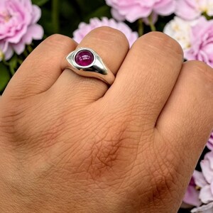 May include: A polished silver ring featuring a smooth, rounded band and a central, bezel-set cabochon gemstone in a deep, vibrant magenta-pink color. The setting has an elongated, eye-like shape that tapers toward the sides of the finger.