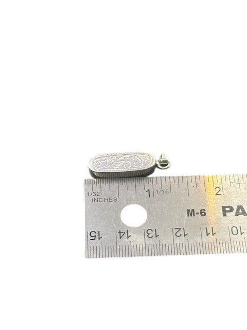 May include: Silver pendant with an engraved floral design. The pendant is oval-shaped and measures approximately 1 inch in length. It has a small loop at the top for a chain. The pendant is shown next to a ruler.
