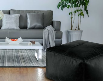 May include: A modern living room scene featuring a gray sofa, white coffee table, and a black square ottoman. A terracotta vase with white flowers sits on the table. A potted plant adds a touch of greenery.