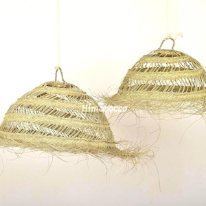 May include: Two pendant lights with a natural, woven design. The lampshades are bell-shaped, made of light beige straw, and have a fringed edge. They hang from thin, off-white cords against a plain background, creating a rustic, bohemian aesthetic.