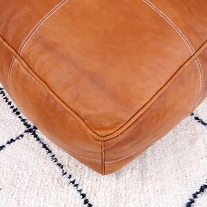 May include: A tan leather ottoman with white stitching. The ottoman is sitting on a white rug with black geometric patterns.