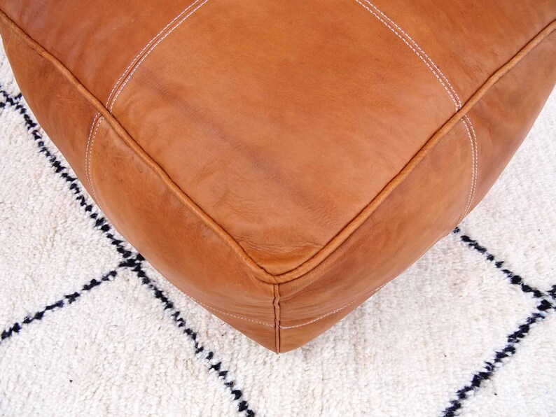 May include: A tan leather ottoman with white stitching. The ottoman is sitting on a white rug with black geometric patterns.