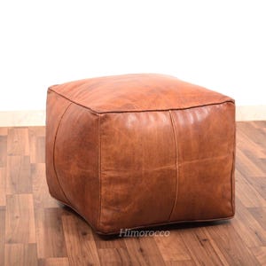 May include: A brown leather pouf ottoman with a square shape. The pouf is made of thick, durable leather and has a simple, elegant design.