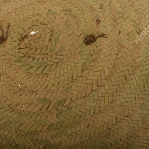 K&ouml;nnte beinhalten: Nahaufnahme eines geflochtenen Strohhutes. Der Hut besteht aus naturfarbenem Stroh mit einem dicht gewebten Muster. Der Rand ist leicht gebogen und die Krone hat eine abgerundete Form. Der Hut hat zwei kleine, dunkel gef&auml;rbte Bindeb&auml;nder.