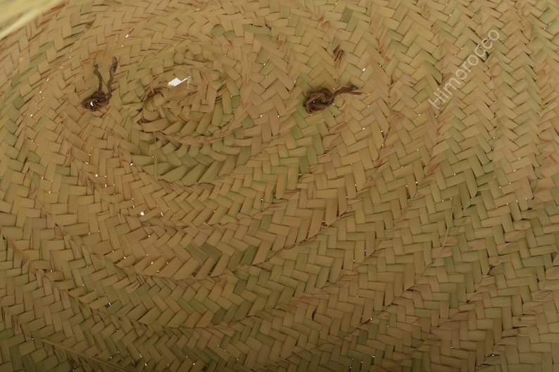 K&ouml;nnte beinhalten: Nahaufnahme eines geflochtenen Strohhutes. Der Hut besteht aus naturfarbenem Stroh mit einem dicht gewebten Muster. Der Rand ist leicht gebogen und die Krone hat eine abgerundete Form. Der Hut hat zwei kleine, dunkel gef&auml;rbte Bindeb&auml;nder.