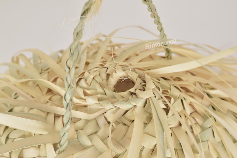 K&ouml;nnte beinhalten: Nahaufnahme eines Lampenschirms aus gewebten Naturfasern. Der Lampenschirm besteht aus hellbeige gewebten Streifen mit einer geflochtenen Aufh&auml;ngeschnur. Das Gewebe erzeugt ein strukturiertes, offenes Design. Die Gesamt&auml;sthetik ist rustikal und organisch.