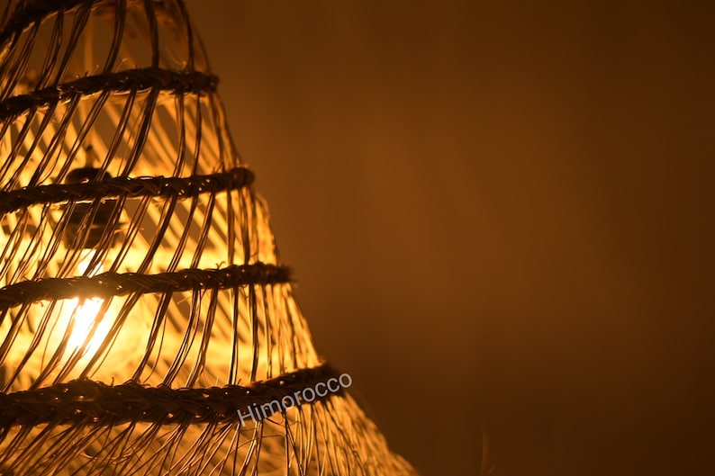 May include: A woven lampshade made of natural materials, with a warm, glowing light emanating from within. The lampshade is cone-shaped and has a rustic, handcrafted look.
