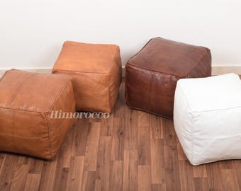 May include: Four square leather poufs in brown, tan, and white. The poufs are made of leather and have a square shape. They are sitting on a wooden floor.