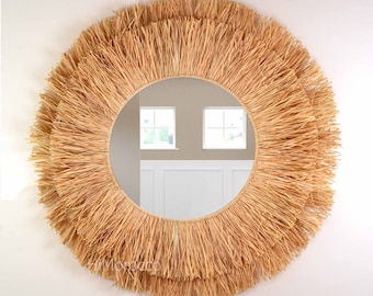 May include: A round mirror with a natural woven straw frame. The frame has a thick, layered fringe that creates a textured look. The mirror reflects a room with two windows.