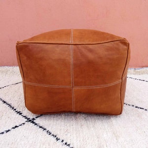 May include: A brown leather pouf with white stitching. The pouf is sitting on a white rug with black diamond patterns.