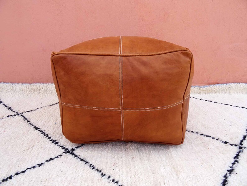 May include: A brown leather pouf with white stitching. The pouf is sitting on a white rug with black diamond patterns.
