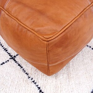 May include: A brown leather ottoman with white stitching. The ottoman is sitting on a white rug with black geometric patterns.