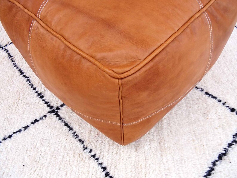 May include: A brown leather ottoman with white stitching. The ottoman is sitting on a white rug with black geometric patterns.