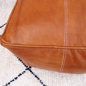 May include: A brown leather pouf with white stitching. The pouf is sitting on a white and black rug.