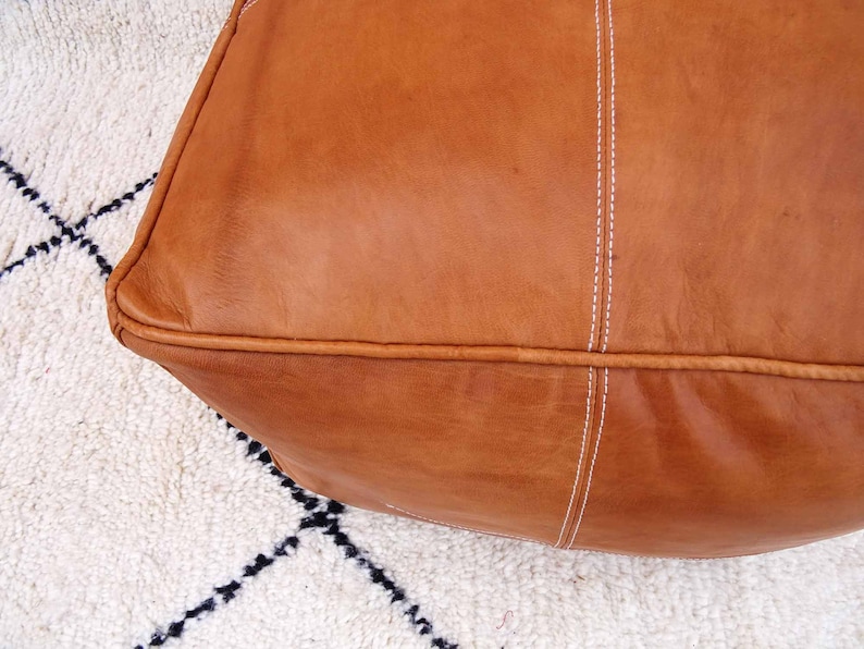 May include: A brown leather pouf with white stitching. The pouf is sitting on a white and black rug.