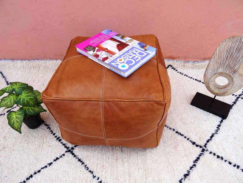 May include: A brown leather pouf with white stitching sits on a white rug with black geometric patterns. A book titled "DECO MAISON" is on top of the pouf.