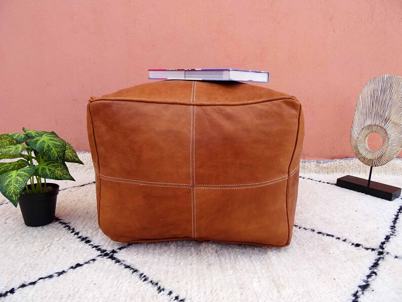 May include: A brown leather pouf with white stitching. The pouf is sitting on a white rug with black squares. A book is sitting on top of the pouf.