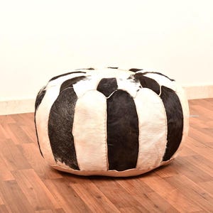 Könnte beinhalten: Ein runder, schwarz-weiß gestreifter Ottoman. Der Pouf besteht aus einem strukturierten Material und steht auf einem Holzboden. Die schwarzen Streifen sind vertikal und wechseln sich mit weißen Streifen ab.