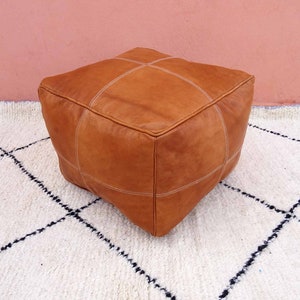 May include: A brown leather ottoman with white stitching. The ottoman is sitting on a white rug with black diamond patterns.