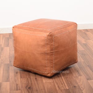 May include: A square brown leather ottoman with white stitching. The ottoman is sitting on a wooden floor.