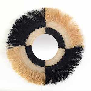 May include: A round mirror with a black and natural woven frame. The frame has a fringe border.