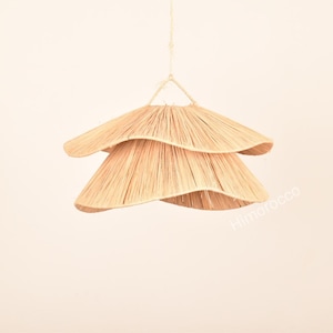 May include: A layered pendant light made from natural fibres, resembling a flower. The light fitting is suspended by a rope and has two tiers of woven material in a light beige colour. The design is organic and minimalist.