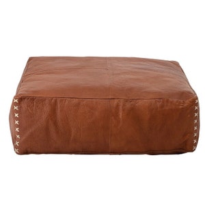 May include: A brown leather ottoman with white stitching around the edges. The ottoman is square and has a tufted top.