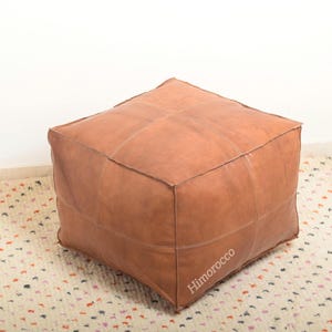 May include: A square brown leather ottoman with a stitched design. The ottoman is sitting on a white rug with small colorful dots.