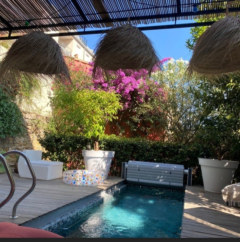 K&ouml;nnte beinhalten: Terrasse im Freien mit kleinem Swimmingpool, umgeben von &uuml;ppigem Gr&uuml;n und leuchtenden Blumen. Der Bereich ist von einem gewebten Baldachin beschattet und verf&uuml;gt &uuml;ber dekorative H&auml;ngelampen. Wei&szlig;e Pflanzgef&auml;&szlig;e und Sitzgelegenheiten tragen zur entspannten Atmosph&auml;re bei.