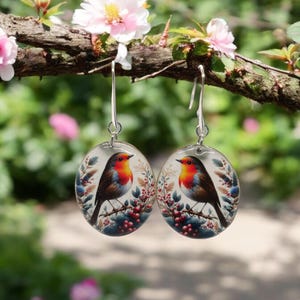 May include: A pair of silver dangle earrings with a round, clear glass charm featuring a robin perched on a branch with red berries. The robin has a red breast, brown wings, and a white belly.