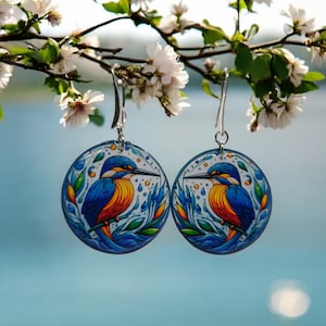 May include: A pair of round earrings with a colorful illustration of a kingfisher bird on a blue background. The earrings are silver and have a hook closure.