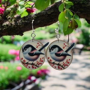 May include: A pair of silver earrings with round, white, and red painted wood pendants featuring a black and white bird perched on a branch with red berries.