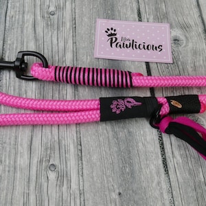 Könnte beinhalten: Rosa-schwarzes Hundeleine aus Seil mit schwarzem Griff und rosa-schwarzem Quasten. Die Leine hat einen schwarzen Metallclip und ein schwarzes Schild mit dem Text "Live Pawlicious".