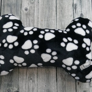 Op de afbeelding: Zwart-witte pluche hondenbotvormig kussen met een pootprintpatroon. Het kussen heeft een wit label met het woord "Pawlicious" erop.