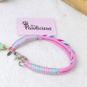 Op de afbeelding: Een roze en turquoise hondenriem met een gevlochten gedeelte en een kwastje. De halsband heeft een zilveren sluiting en een label. De merknaam "Liba Pawlicious" staat op een roze kaart.