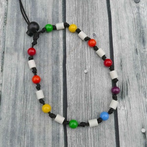 Könnte beinhalten: Eine schwarze Schnurkette mit weißen, roten, gelben, grünen, blauen und violetten Perlen. Die Perlen sind in einem Regenbogenmuster angeordnet.