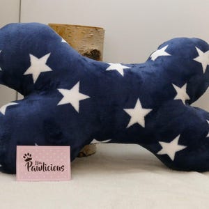 Op de afbeelding: Een marineblauw, botvormig kussen voor huisdieren met witte sterpatronen. Het kussen is gemaakt van zachte stof en is ontworpen voor huisdieren. Een kleine roze kaart met de tekst "Pawlicious" staat voor het kussen.