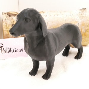 Op de afbeelding: Zwarte teckelhond figuur. Het beeldje is gemaakt van glad, donkergrijs materiaal en staat op een wit oppervlak.