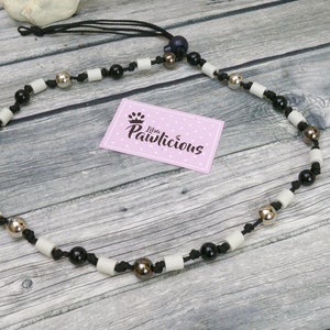 Op de afbeelding: Een zwart-witte kralenketting met zilveren en gouden kralen. De ketting is gemaakt met een zwart koord en heeft een klein zwart label met de tekst "Little Pawlicious" erop.