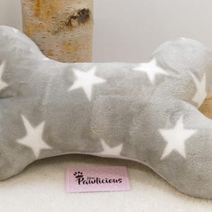 Op de afbeelding: Een grijs, botvormig hondenbed met witte sterpatronen. Het pluche bed heeft een zachte textuur en is ontworpen voor het comfort van huisdieren. Een klein roze label met de tekst "Pawlicious" is zichtbaar.