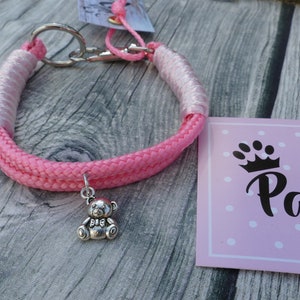 Op de afbeelding: Roze en witte touwhalster met een zilveren berenbedel. Het halster heeft een zilveren sluiting en is aan de ene kant versierd met een wit touw en aan de andere kant met een roze touw. De bedel is met een zilveren ring aan het halster bevestigd. Het halster ligt op een houten oppervlak. De tekst "Liba Pawlicia" staat op een roze kaart met witte stippen.