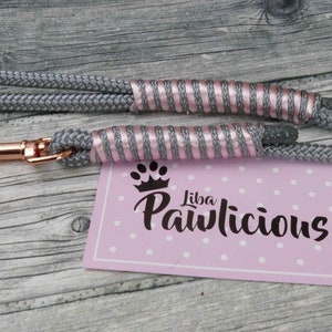 Könnte beinhalten: Eine graue Hundeleine aus Seil mit einem roségoldenen Haken und rosa-silbernen Streifen. Die Leine liegt auf einer Holzoberfläche mit einer rosa Karte, auf der "Liba Pawlicious" mit einem Pfotenabdruck und einer Krone steht.