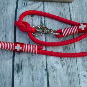 Op de afbeelding: Rode touwhalster voor honden met een wit en rood Zwitsers kruisontwerp. De halster heeft een zilveren metalen clip.