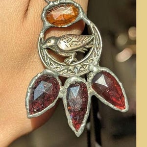 May include: A silver-toned metal pendant featuring a central circular frame with a bird relief and intricate knotwork. The pendant is adorned with one oval amber-colored stone at the top and three faceted, teardrop-shaped dark red stones below.