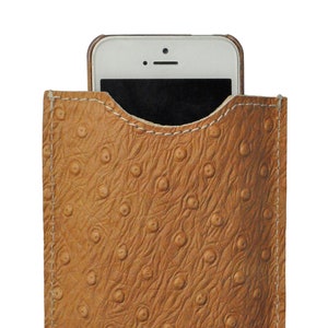 Puede incluir: Una funda de teléfono de cuero marrón con un patrón de piel de avestruz. La funda tiene un detalle de costura blanca alrededor de los bordes.