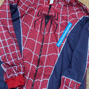 May include: A red and blue zip-up hooded sweatshirt featuring a black spider emblem on the chest and a white web pattern across the red sections. The garment includes a black zip, red ribbed cuffs, a red hem, and a blue label reading PeterTechIndustries.
