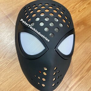 Może przedstawiać: Czarna plastikowa maska Spider-Mana z logo PeterTechIndustries i białym siateczkowym okiem.