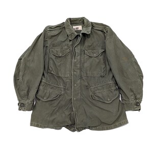 May include: Olive green military-style jacket with four front pockets and button closures. The jacket has a collared neckline and buttoned cuffs. The fabric appears worn, suggesting a vintage or used condition. The jacket is laid flat against a white background.